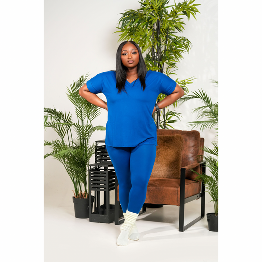 Woman in a blue outfit standing in a room with plants and a brown chair.