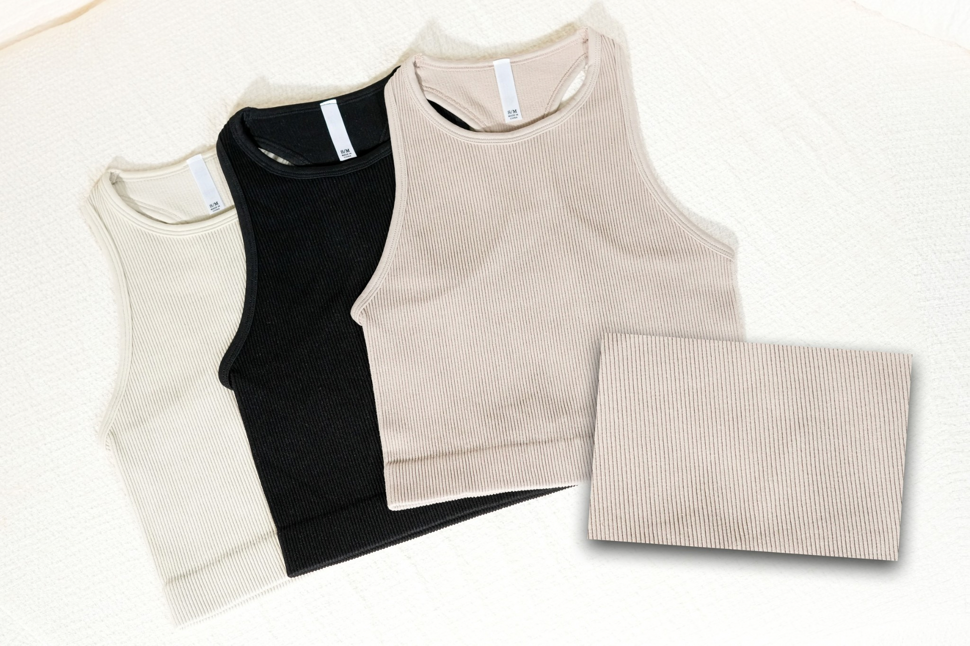 Three tank tops in beige, black, and beige with a folded beige fabric on a white background. RIBBED SEAMLESS HIGH-NECK CROPPED TANK TOP. BLACK, mocha, sand beige